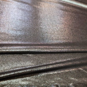 May include: Close-up of a metallic fabric with a subtle iridescent sheen. The fabric has a textured surface and is folded in a few places.