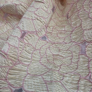 May include: Close-up of a cream fabric with a floral pattern. The design features large, stylized flowers outlined in pink. The fabric has a textured, slightly wrinkled appearance, with folds and shadows adding depth to the image. The overall effect is elegant and delicate.
