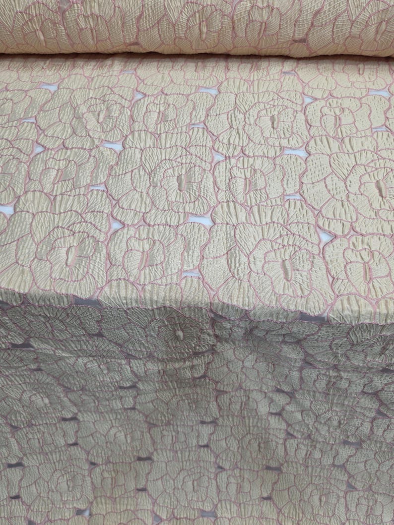 May include: Close-up of a fabric roll with a repeating floral pattern. The fabric has a textured, quilted appearance with a light beige base and delicate pink outlines. The floral design is evenly spaced across the material.