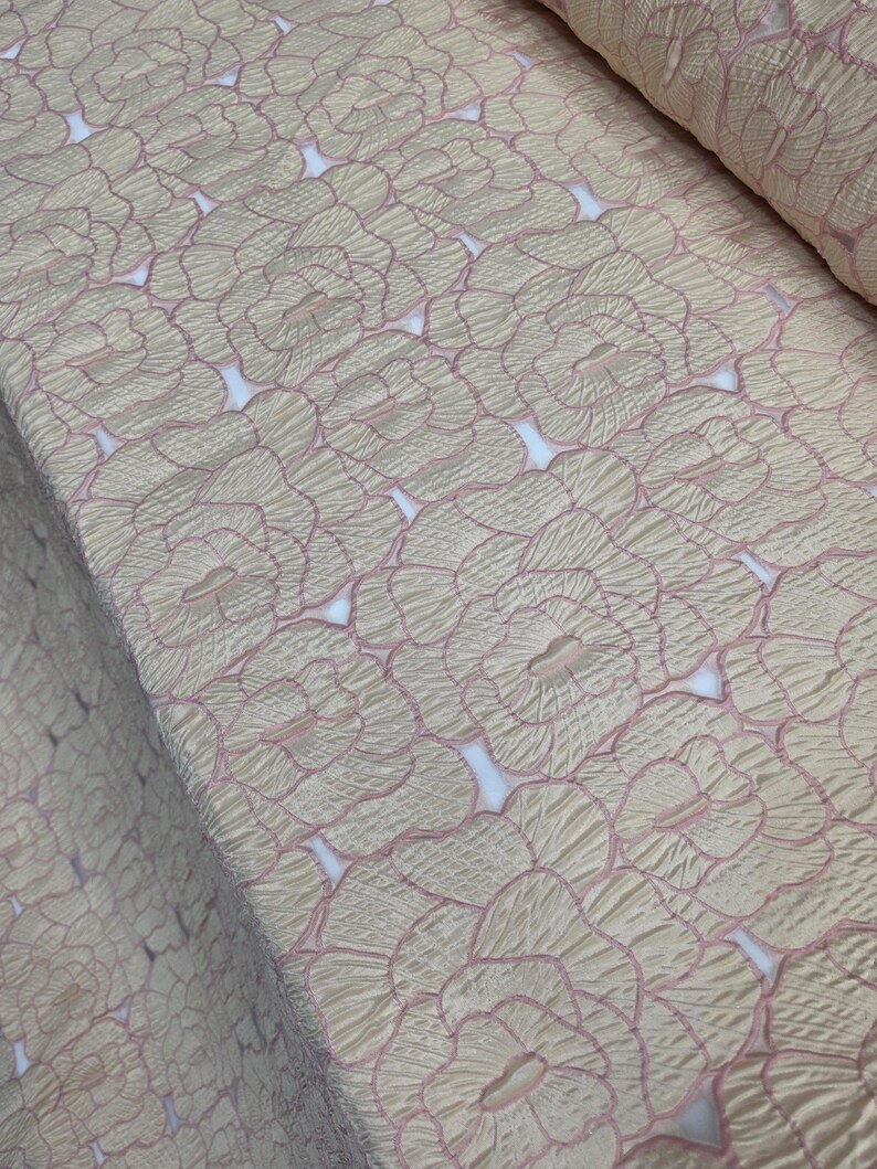 May include: Close-up of a fabric with a repeating floral pattern. The fabric has a textured surface with a light beige background and pink outlines. The floral design is visible throughout the fabric.