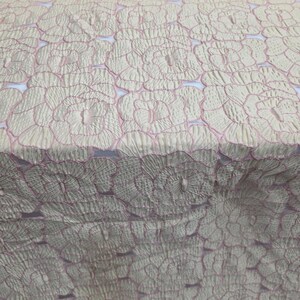 May include: Pale cream fabric featuring a repeating floral design with pink outlines. The material has a textured appearance, possibly quilted or embroidered. The floral pattern is consistently spaced, giving a delicate and elegant aesthetic.