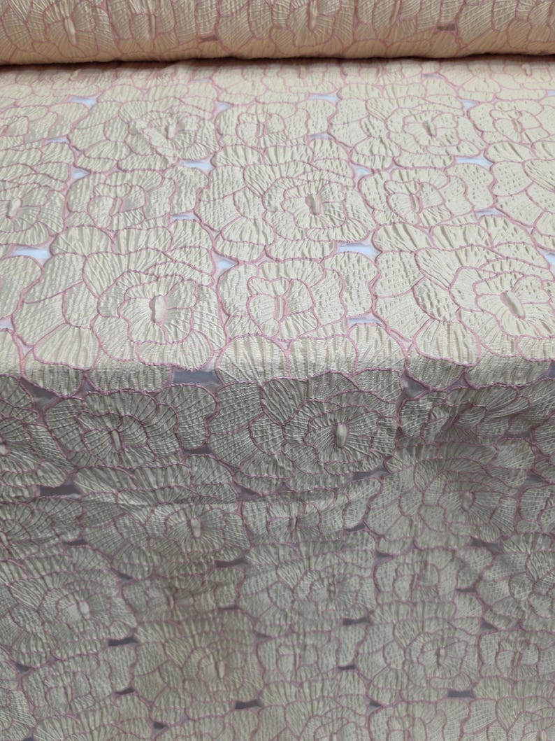 May include: Pale cream fabric featuring a repeating floral design with pink outlines. The material has a textured appearance, possibly quilted or embroidered. The floral pattern is consistently spaced, giving a delicate and elegant aesthetic.