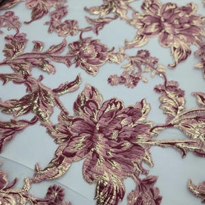May include: Close-up of a fabric with a floral design. The fabric features large, detailed flowers and leaves in shades of mauve and gold. The gold accents add a touch of elegance to the design. The fabric appears to be a delicate material, possibly for special occasion wear.