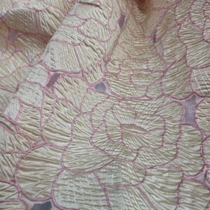 May include: Close-up of a cream fabric with a floral pattern. The design features large, stylized flowers outlined in pink. The fabric has a textured, slightly wrinkled appearance, with folds and shadows adding depth to the image. The overall effect is elegant and delicate.