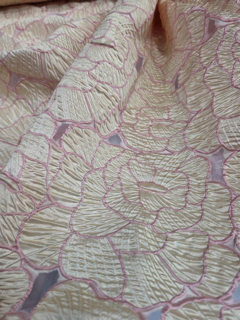 May include: Close-up of a cream fabric with a floral pattern. The design features large, stylized flowers outlined in pink. The fabric has a textured, slightly wrinkled appearance, with folds and shadows adding depth to the image. The overall effect is elegant and delicate.