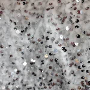 May include: Close-up of a white fabric covered in silver sequins. The sequins are round and reflect light, creating a shimmering effect. The fabric appears to be a decorative textile, possibly for clothing or accessories.