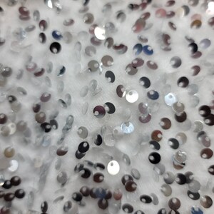May include: Close-up of a white fabric covered in small, silver sequins. The sequins are round and reflective, catching the light to create a shimmering effect. The fabric appears soft and textured, with the sequins scattered across the surface.