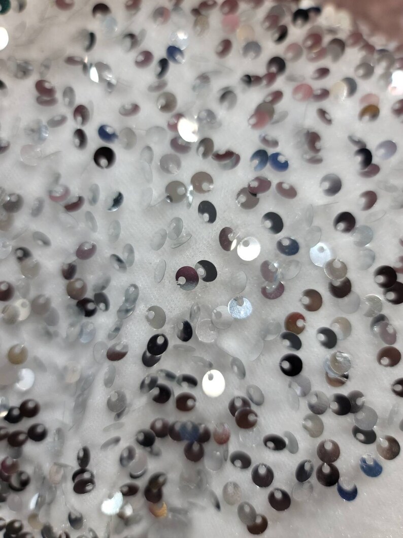 May include: Close-up of a white fabric covered in small, silver sequins. The sequins are round and reflective, catching the light to create a shimmering effect. The fabric appears soft and textured, with the sequins scattered across the surface.