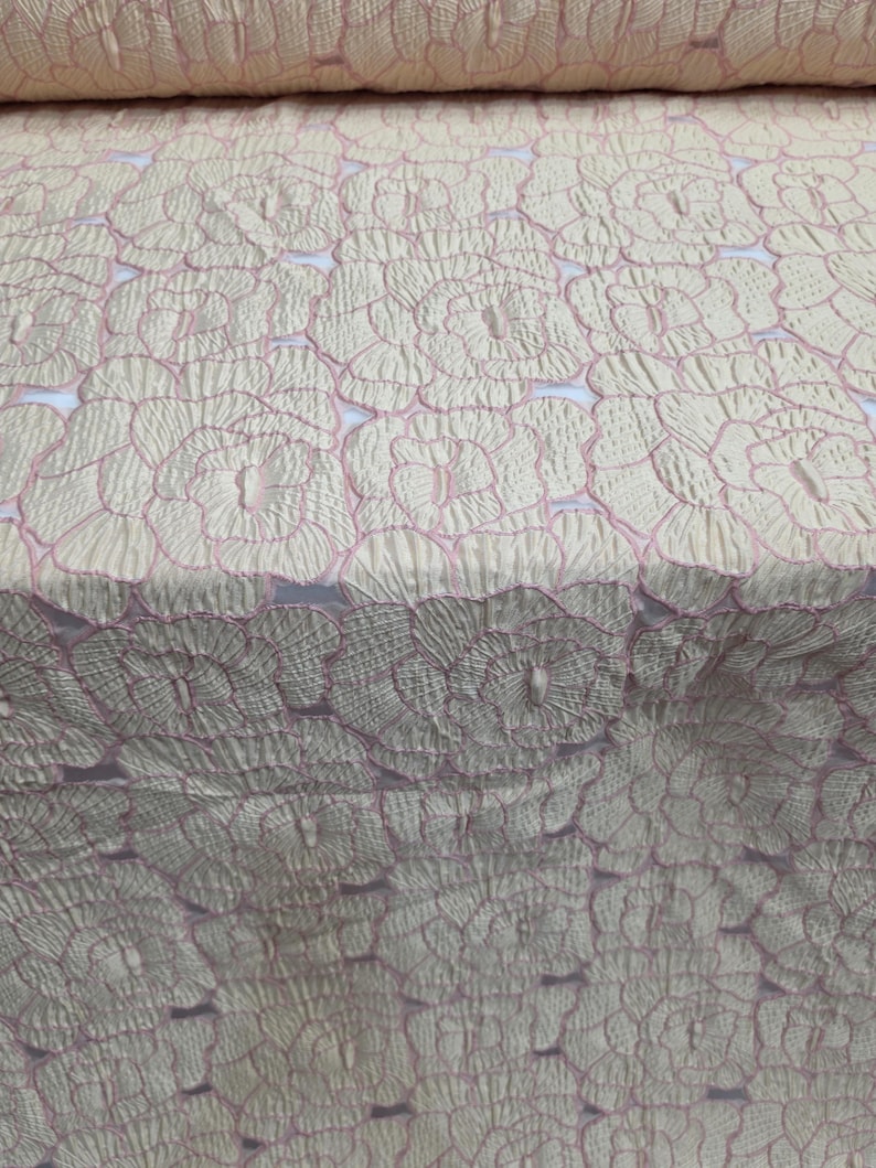 May include: Pale cream fabric featuring a repeating floral design with pink outlines. The material has a textured appearance, possibly quilted or embroidered. The floral pattern is consistently spaced, giving a delicate and elegant aesthetic.