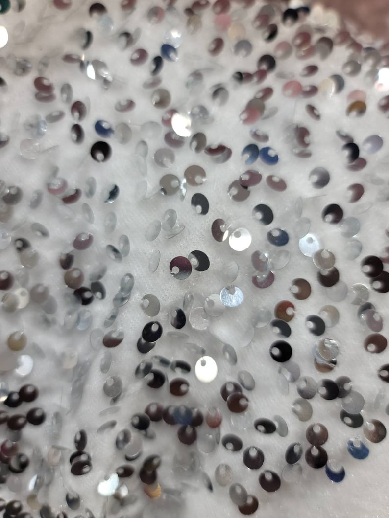 May include: Close-up of a white fabric covered in small, silver sequins. The sequins are round and reflect light, creating a shimmering effect. The fabric appears soft and textured, with the sequins scattered across the surface.