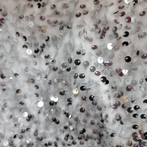 May include: Close-up of a white fabric covered in silver sequins. The sequins are round and reflect light, creating a shimmering effect. The fabric appears to be a delicate material, possibly for a dress or other formal wear.