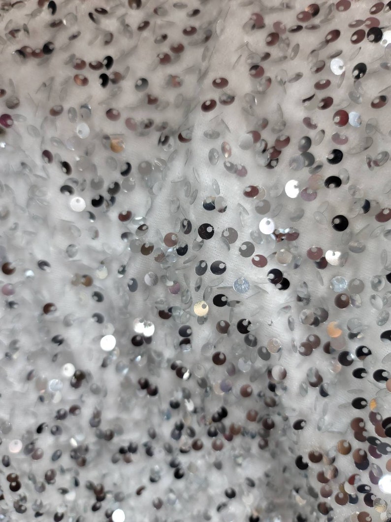 May include: Close-up of a white fabric covered in silver sequins. The sequins are round and reflect light, creating a shimmering effect. The fabric appears to be a delicate material, possibly for a dress or other formal wear.