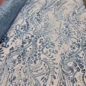 May include: A roll of light blue fabric with intricate, shimmering floral and scrollwork patterns. The fabric is embellished with sparkling sequins, creating a luxurious and elegant appearance. The design is set against a white background.