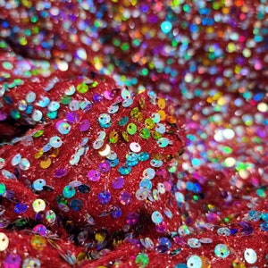 May include: Close-up of red fabric covered in iridescent sequins. The sequins are in various colors, including blue, green, yellow, and purple. The fabric is textured and appears to be folded, creating shadows and highlights.