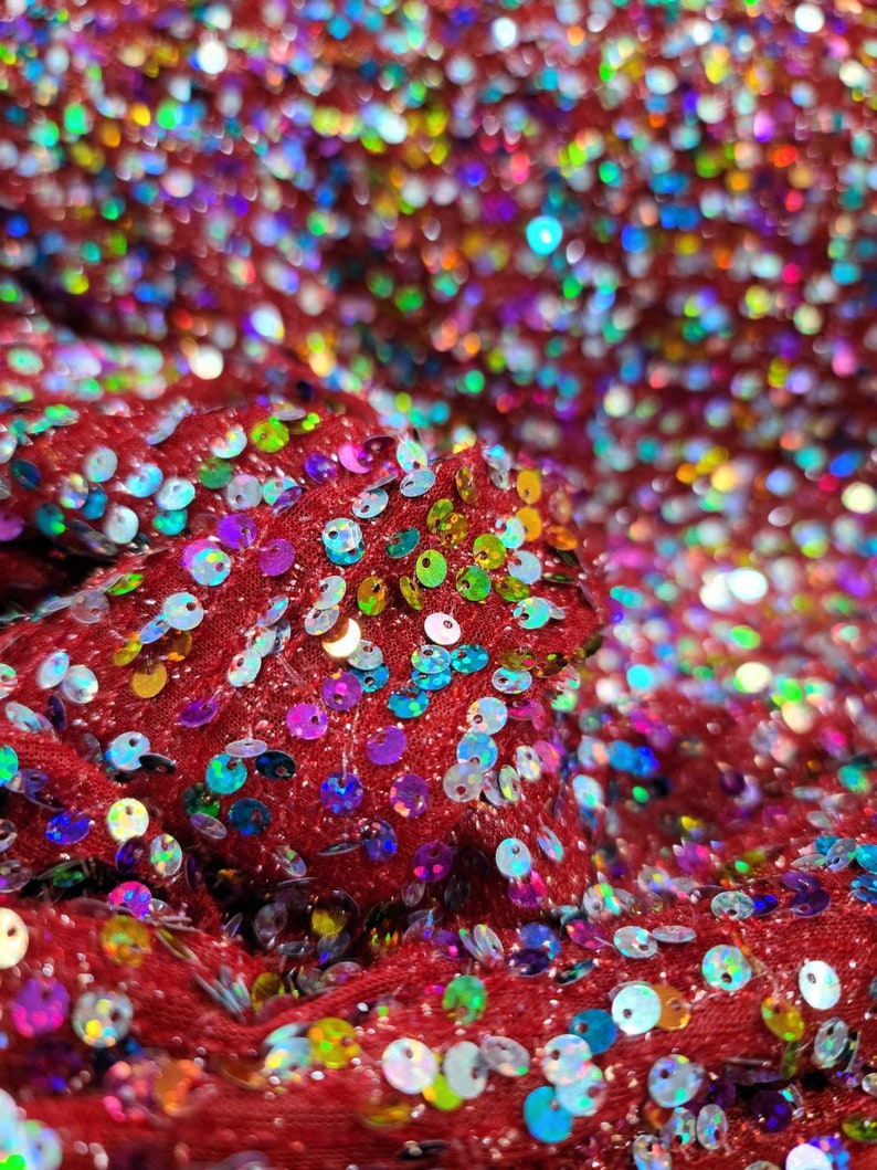 May include: Close-up of red fabric covered in iridescent sequins. The sequins are in various colors, including blue, green, yellow, and purple. The fabric is textured and appears to be folded, creating shadows and highlights.
