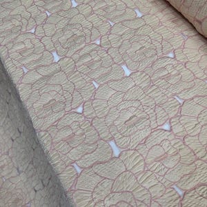 May include: Close-up of a fabric with a repeating floral pattern. The fabric has a textured appearance with a light beige base and pink outlines defining the flower shapes. The design creates a sense of depth and dimension.