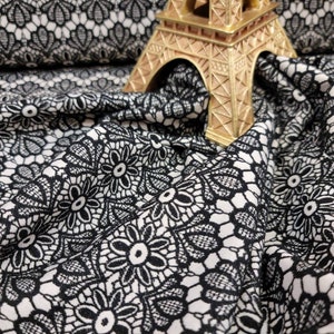 May include: Black and white floral lace fabric with a repeating pattern. A gold Eiffel Tower figurine sits atop the fabric. The fabric is draped, showing the texture and design of the lace.