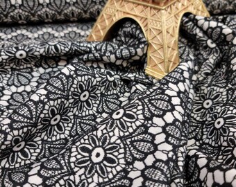 May include: Black and white floral lace fabric with a repeating pattern. A gold Eiffel Tower figurine sits atop the fabric. The fabric is draped, showing the texture and design of the lace.