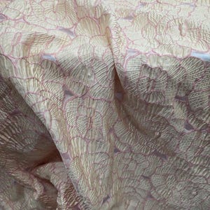 May include: Close-up of a fabric with a floral pattern. The fabric has a textured, shimmering appearance with a light peach base and pink floral outlines. The design features repeating circular flower shapes, creating a delicate and elegant look.