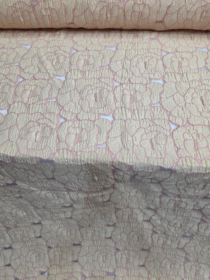 May include: Close-up of a fabric roll with a repeating floral pattern. The fabric has a textured, quilted appearance with a light beige base and delicate pink outlines. The floral design is evenly spaced across the material.