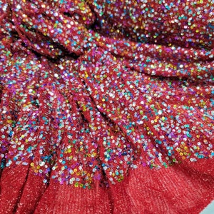 May include: Close-up of a roll of red fabric covered in iridescent sequins. The fabric has a textured, slightly ruffled appearance, with the sequins creating a sparkling effect. The sequins are a mix of colors, including blue, green, and gold.