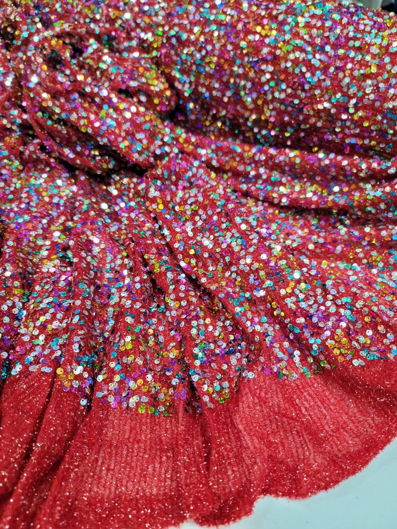 May include: Close-up of a roll of red fabric covered in iridescent sequins. The fabric has a textured, slightly ruffled appearance, with the sequins creating a sparkling effect. The sequins are a mix of colors, including blue, green, and gold.