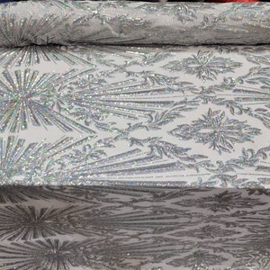 K&ouml;nnte beinhalten: Wei&szlig;er Stoff mit einem silbernen, schimmernden Paillettenmuster. Das Muster zeigt ein sich wiederholendes Design aus stilisierten Blumen und Bl&auml;ttern.
