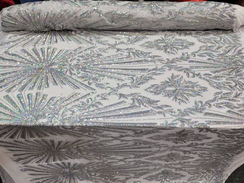 K&ouml;nnte beinhalten: Wei&szlig;er Stoff mit einem silbernen, schimmernden Paillettenmuster. Das Muster zeigt ein sich wiederholendes Design aus stilisierten Blumen und Bl&auml;ttern.