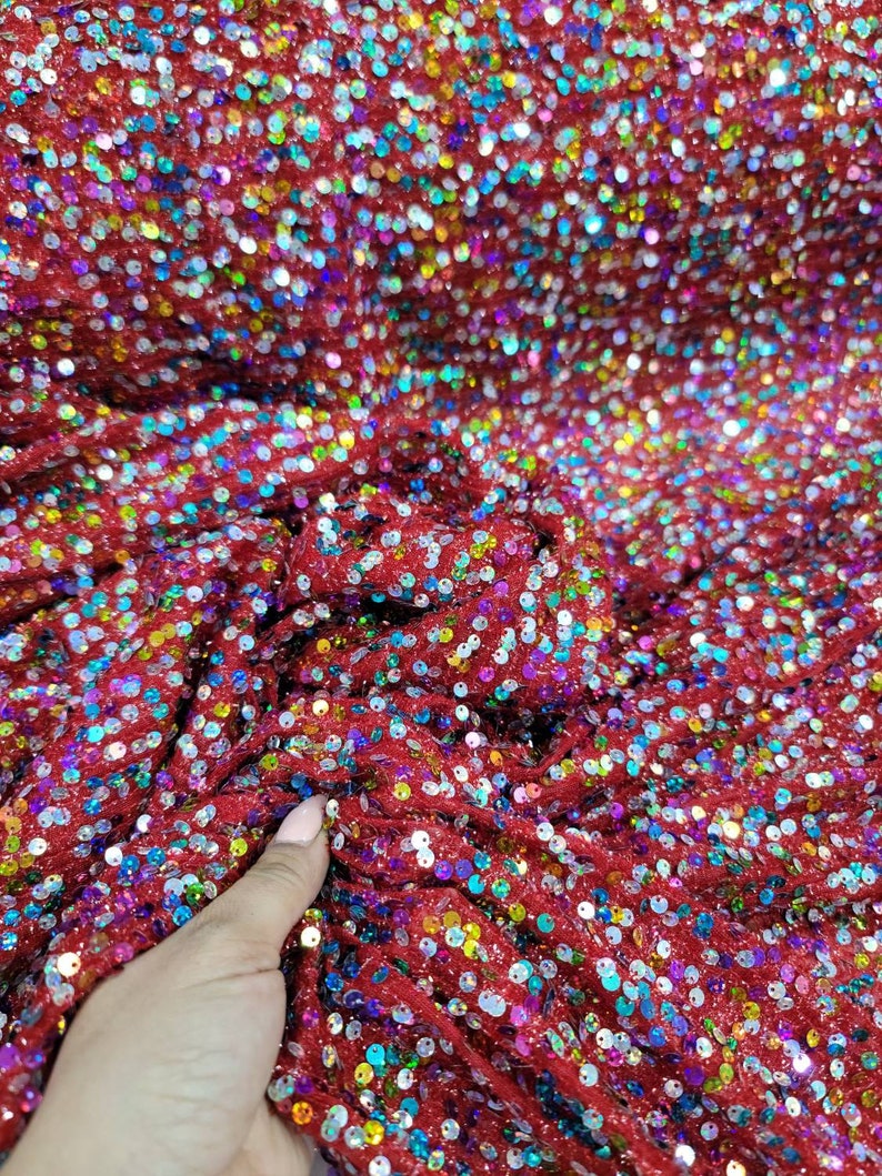 May include: Close-up of red fabric covered in iridescent sequins. The sequins are small and round, reflecting a rainbow of colors. The fabric is slightly ruffled, creating texture and depth. This material is suitable for apparel and craft projects.
