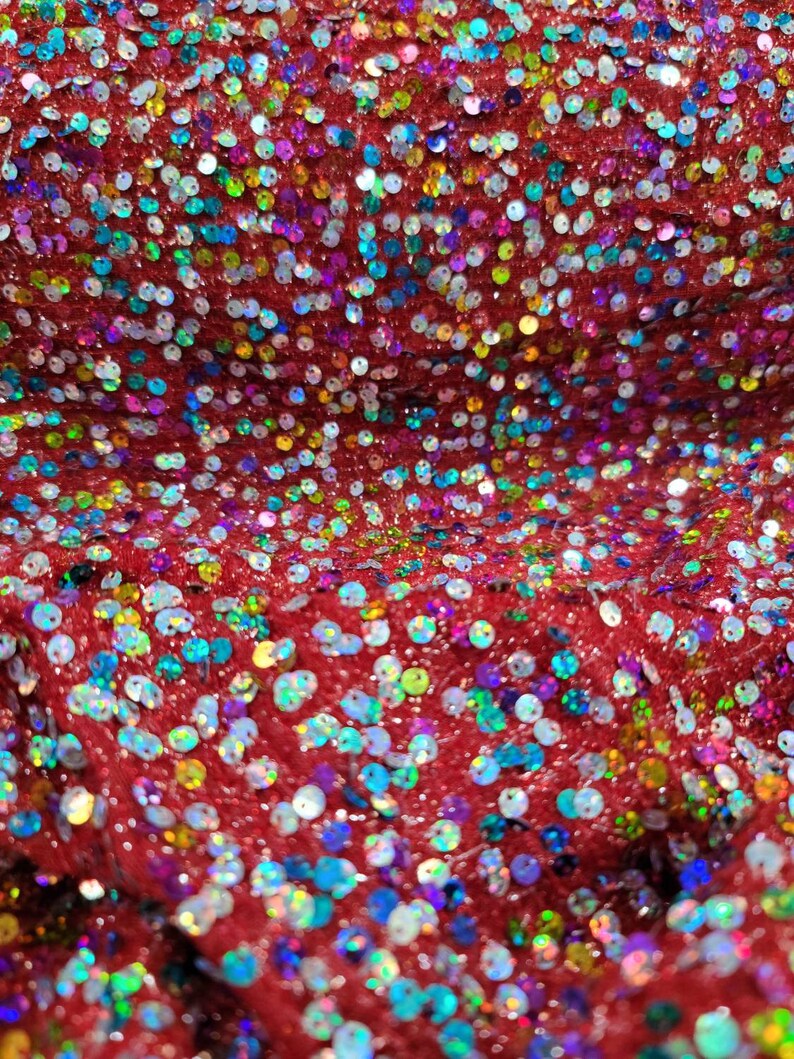 May include: Close-up of red fabric covered in iridescent sequins. The sequins are round and reflect a variety of colors, including blue, green, and purple. The fabric has a textured appearance.