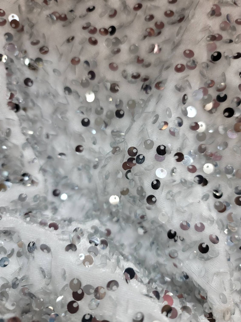 May include: Close-up of a white fabric covered in silver sequins. The sequins are round and vary in size, creating a shimmering effect. The fabric appears soft and textured, with a slight sheen.