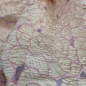 May include: Close-up of a textured fabric with a floral pattern. The fabric is a pale cream colour with pink outlines and accents. The design features overlapping petal-like shapes, creating a layered effect. The fabric appears to be soft and flowing.