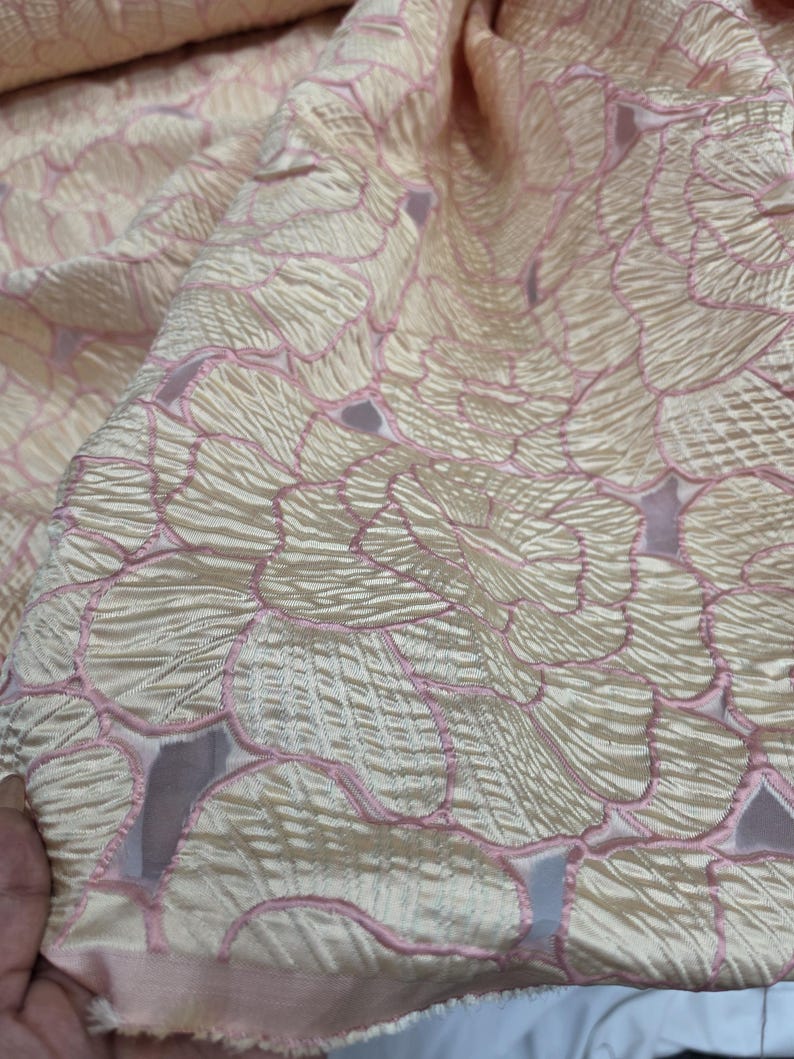 May include: Close-up of a textured fabric with a floral pattern. The fabric is a pale cream colour with pink outlines and accents. The design features overlapping petal-like shapes, creating a layered effect. The fabric appears to be soft and flowing.