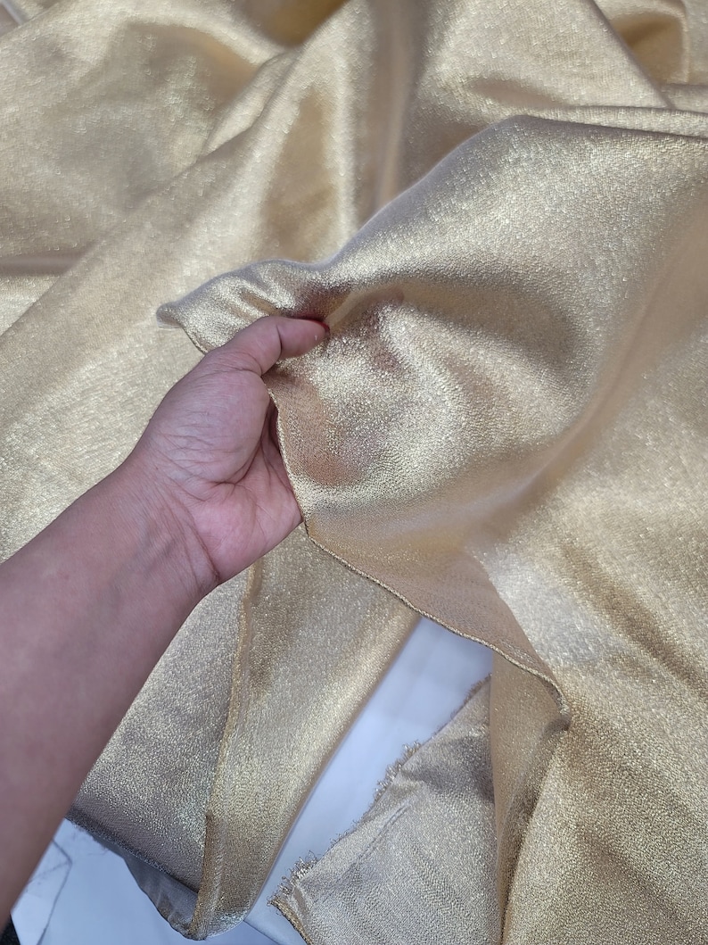 May include: A piece of gold metallic fabric with a textured surface. The fabric is folded and draped over a white surface.