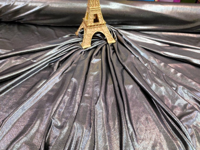 May include: A silver metallic fabric with a subtle iridescent sheen. The fabric is draped and folded, creating a soft, flowing texture.