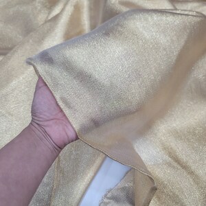 May include: A piece of gold metallic fabric with a shimmery finish. The fabric is folded and held in a hand.