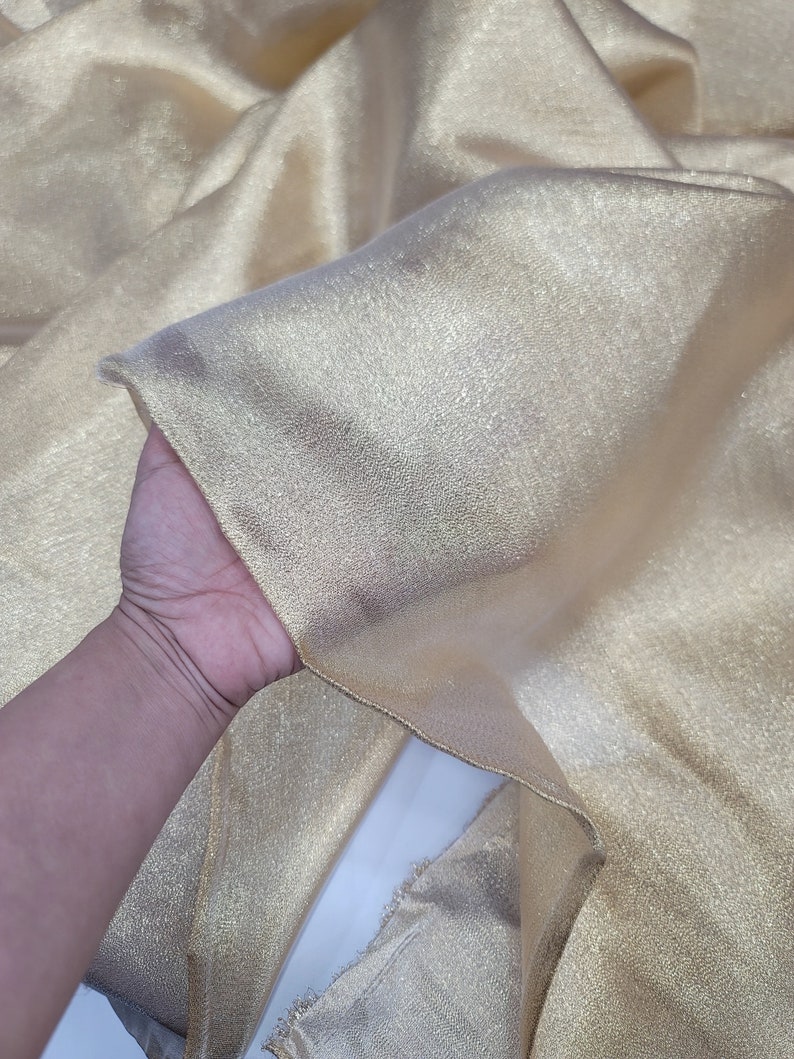 May include: A piece of gold metallic fabric with a shimmery finish. The fabric is folded and held in a hand.