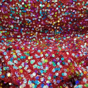 May include: Close-up of red fabric covered in iridescent sequins. The sequins are round and reflect a variety of colors, including blue, green, and purple. The fabric has a textured appearance.