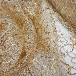 Puede incluir: Tela de encaje con lentejuelas doradas con un patrón delicado e intrincado. La tela es transparente y tiene una textura suave y fluida.