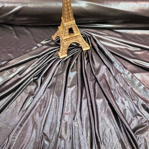 May include: A close-up of iridescent fabric with a metallic sheen. The fabric is draped over itself, creating folds and wrinkles. A small gold Eiffel Tower figurine is placed on top of the fabric.