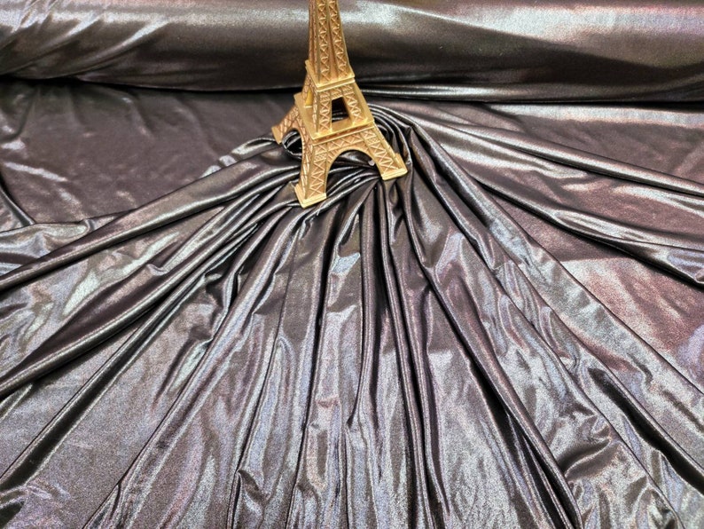 May include: A close-up of iridescent fabric with a metallic sheen. The fabric is draped over itself, creating folds and wrinkles. A small gold Eiffel Tower figurine is placed on top of the fabric.