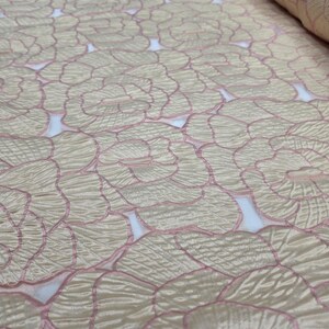 May include: A roll of fabric with a repeating floral design. The fabric has a beige background with pink outlines and details, creating a textured, layered effect. The design is reminiscent of overlapping flower petals.