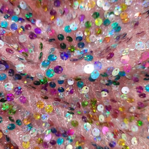 May include: Close-up of pink fabric covered in iridescent sequins. The sequins are in various colors, including blue, green, yellow, purple, and pink. The fabric is slightly wrinkled, creating shadows and depth.