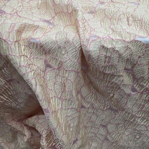 May include: Close-up of a fabric with a floral pattern. The fabric has a textured, shimmering appearance with a light peach base and pink floral outlines. The design features repeating circular flower shapes, creating a delicate and elegant look.