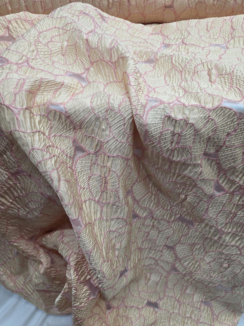 May include: Close-up of a fabric with a floral pattern. The fabric has a textured, shimmering appearance with a light peach base and pink floral outlines. The design features repeating circular flower shapes, creating a delicate and elegant look.