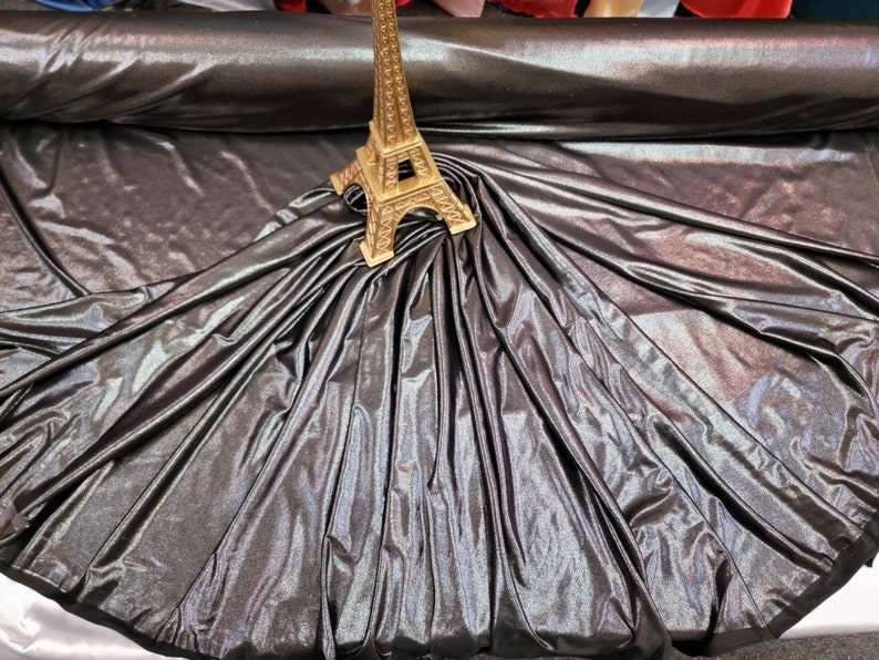 May include: A close-up of iridescent black fabric with a gold Eiffel Tower figurine on top. The fabric is draped and folded, creating a soft, flowing look.