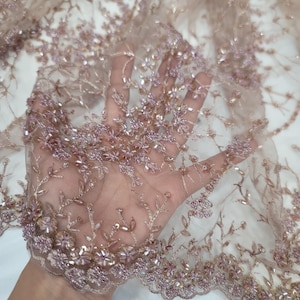 May include: Close-up of a delicate, beaded lace fabric in a soft pink hue. The fabric features a floral pattern with small, iridescent beads and a scalloped edge.