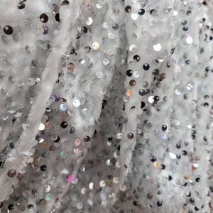 May include: Close-up of a white fabric covered in small, shiny silver sequins. The sequins are round and densely packed, creating a shimmering effect. The fabric has a soft, textured appearance, with visible folds and creases.