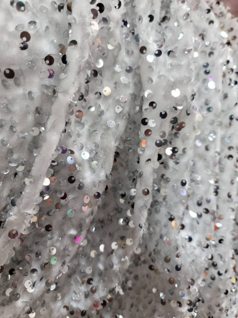 May include: Close-up of a white fabric covered in small, shiny silver sequins. The sequins are round and densely packed, creating a shimmering effect. The fabric has a soft, textured appearance, with visible folds and creases.