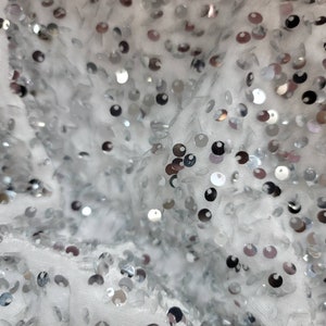 May include: Close-up of a white fabric covered in silver sequins. The sequins are round and vary in size, creating a shimmering effect. The fabric has a soft, textured appearance, with the sequins scattered across the surface.